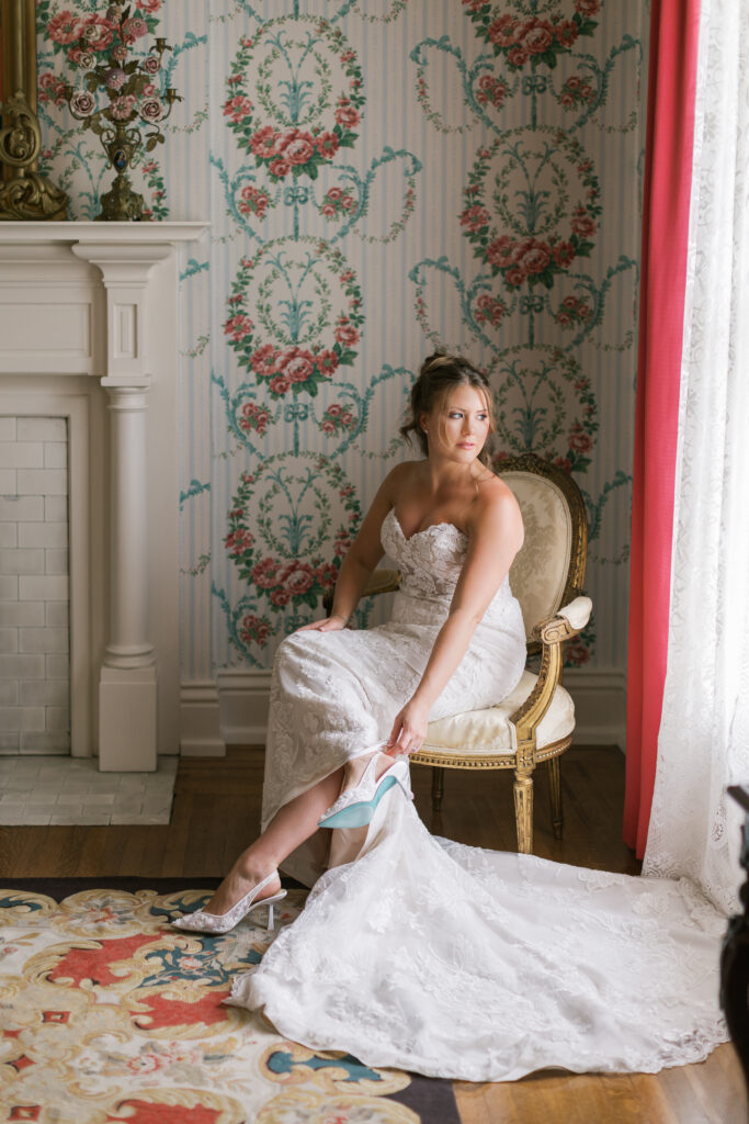 A bride getting ready at Whitehall. She is setting down and putting on her wedding shoes as she looks out the window. 